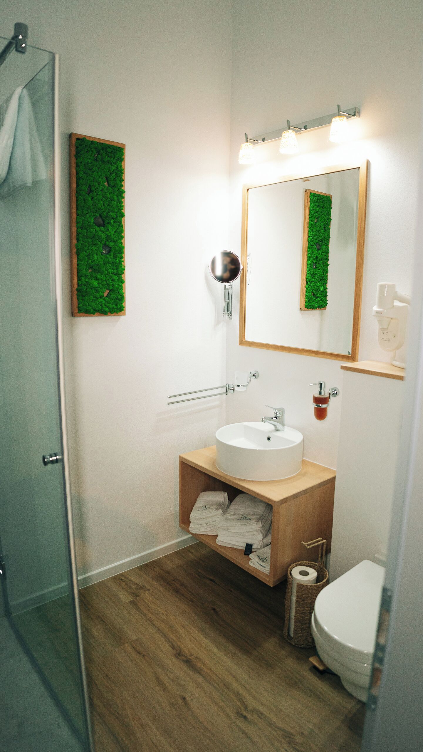 A bathroom with a wooden vanity, a round mirror, a wall-mounted towel rack, a toilet, and a shower area.