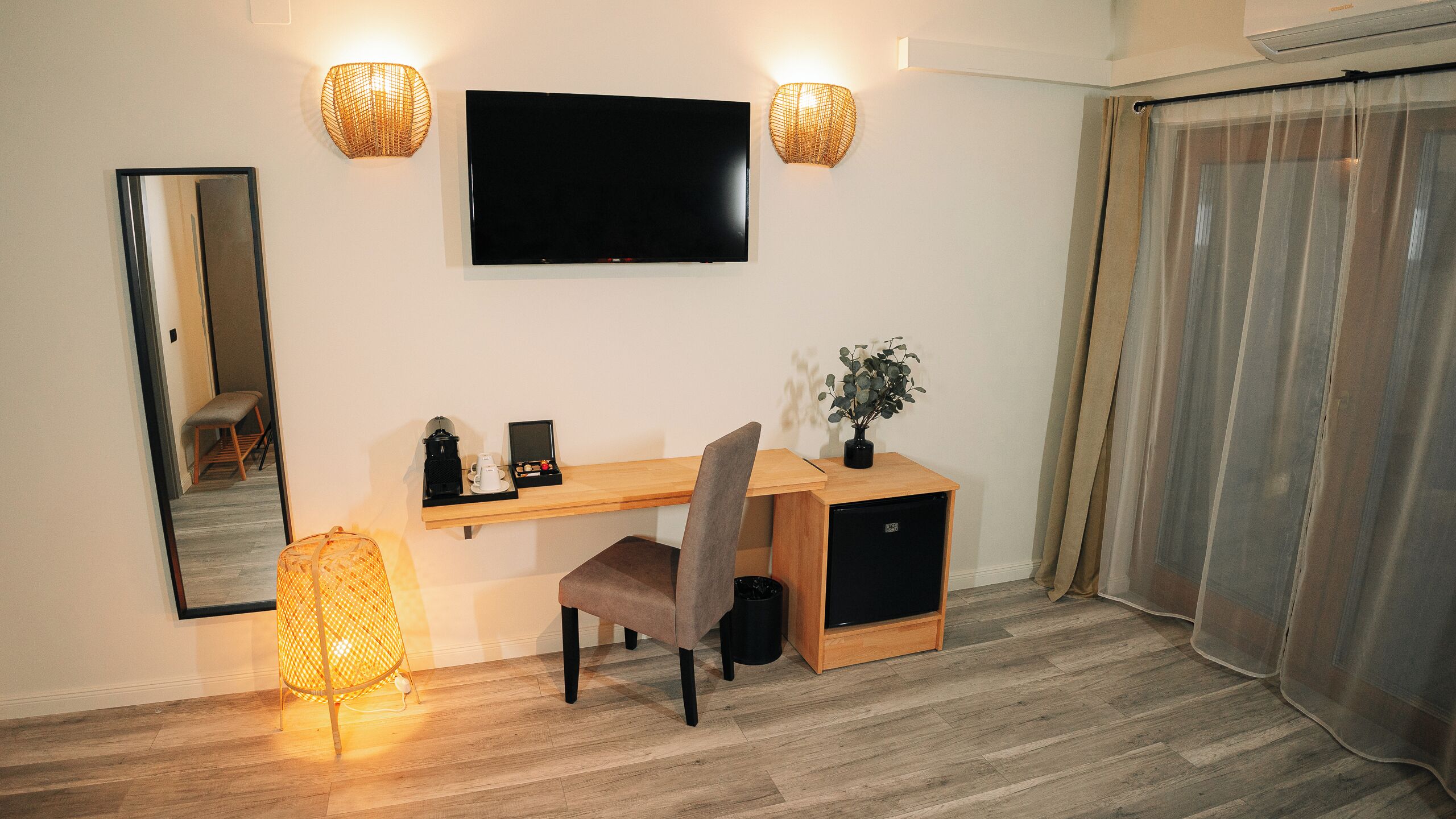 Ett hotellrum med ett skrivbord i trä, en stol, en platt-TV som är monterad på väggen, två vägglampor, en krukväxt och en skjutdörr med gardiner.