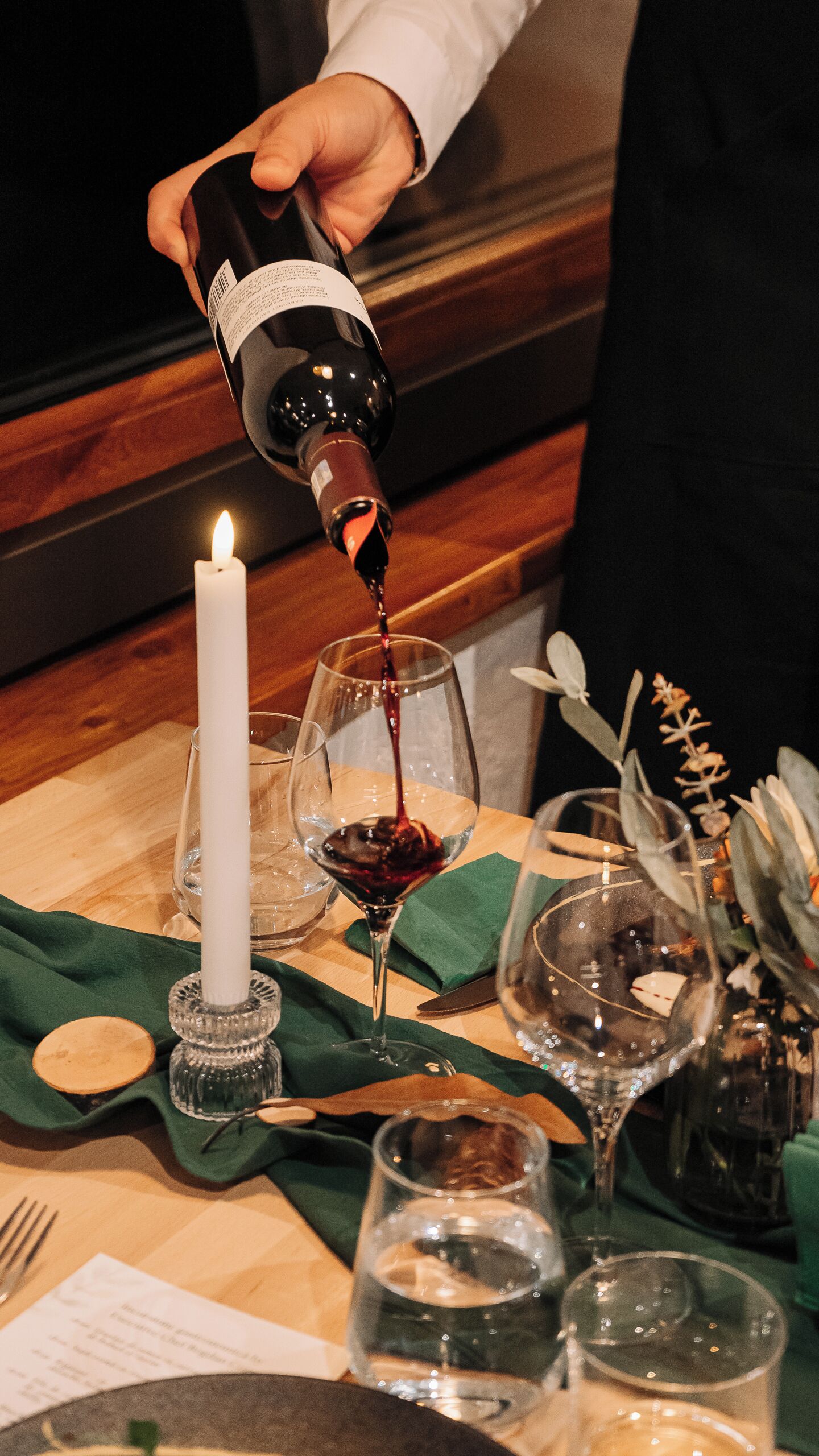 Eine Person gießt Wein ein, eine brennende Kerze und ein Tisch gedeckt mit unterschiedlichem Glas und einer grünen Serviette.
