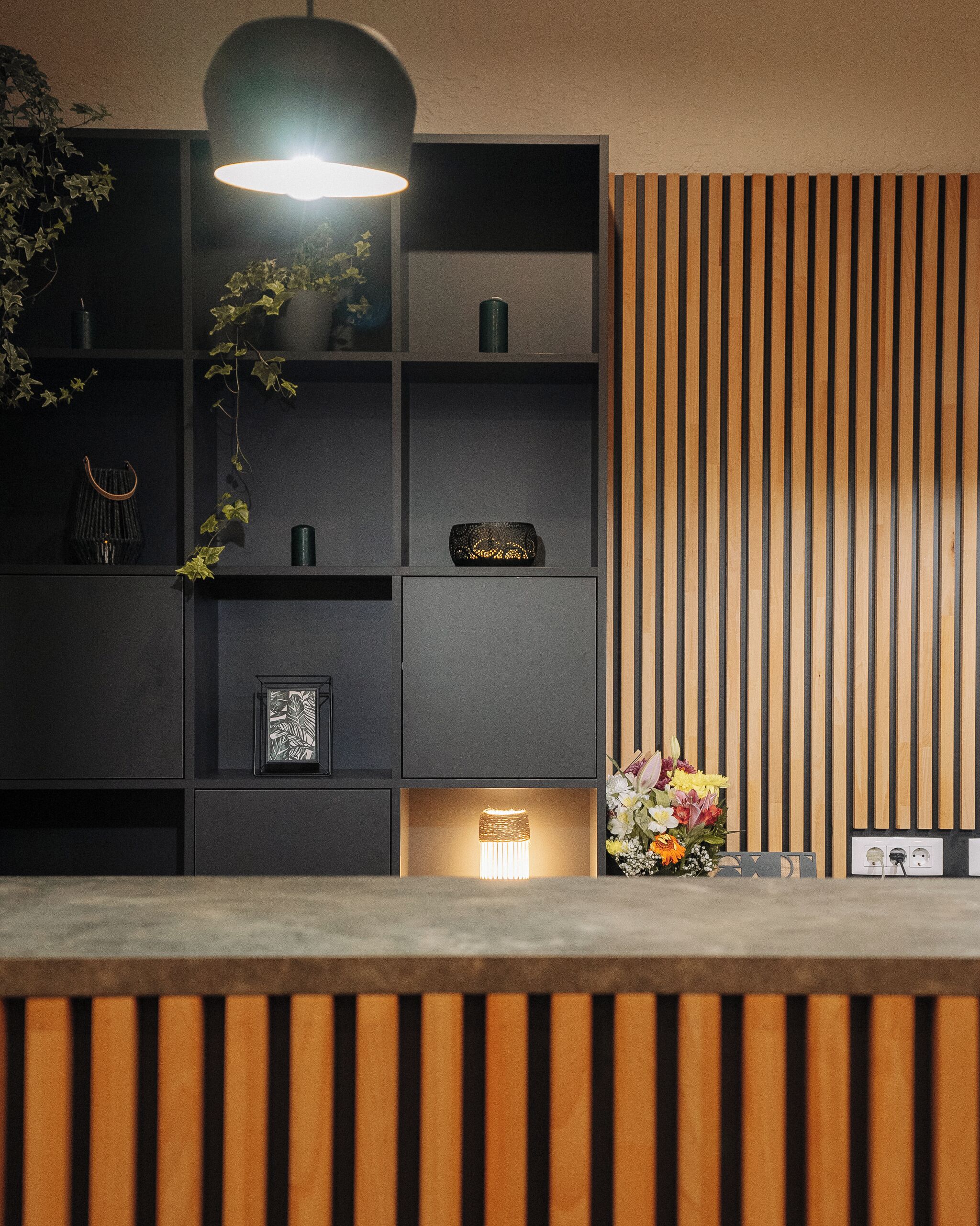 A reception area with a wooden reception desk, a black shelving unit with various items, a hanging light fixture, and a wooden slat wall.