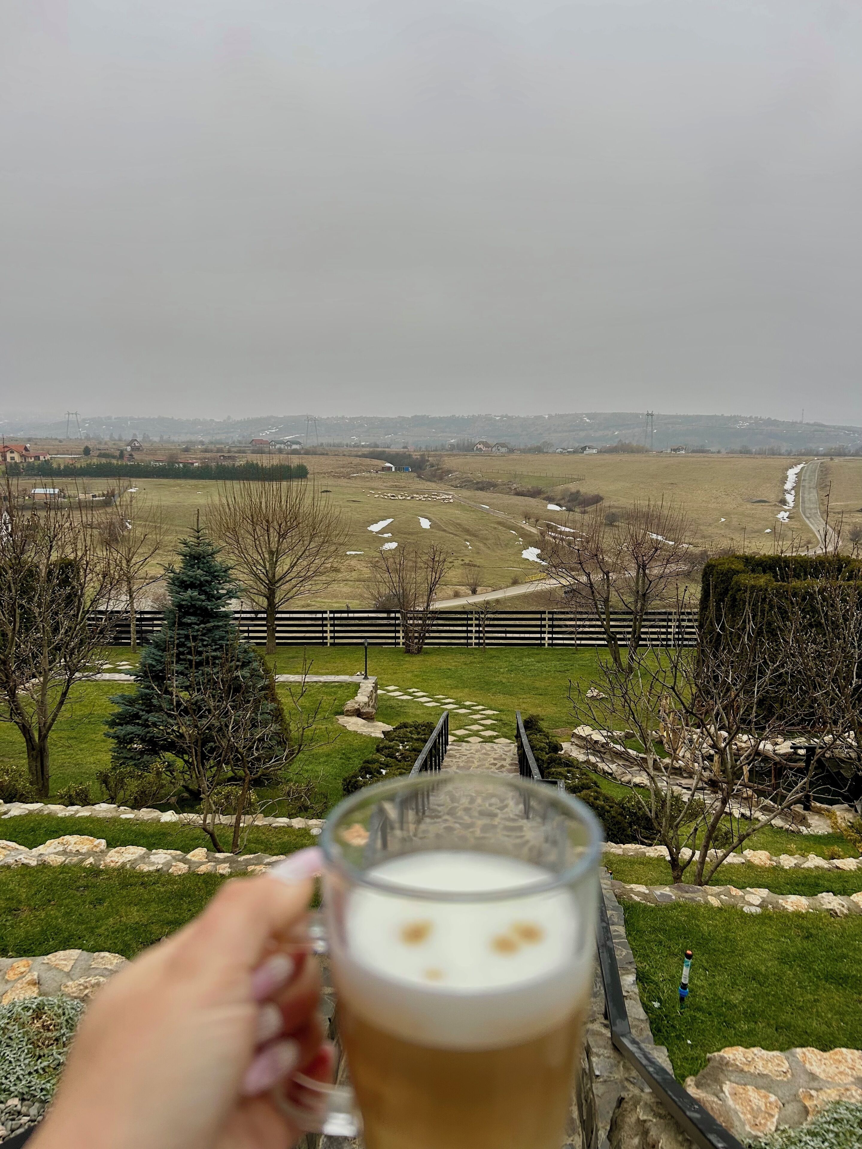 Ein Bierglas vor einer malerischen Aussicht auf eine ländliche Landschaft mit Feldern, einem Zaun und einer in der Ferne liegenden Stadt.