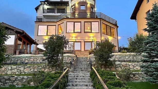 A multi-story building with a stone staircase leading to the entrance, surrounded by greenery. - Nature Nest (Bran)