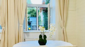 Una habitación con una mesa, dos sillas, un ventanal con cortinas y una planta en maceta con flores blancas.