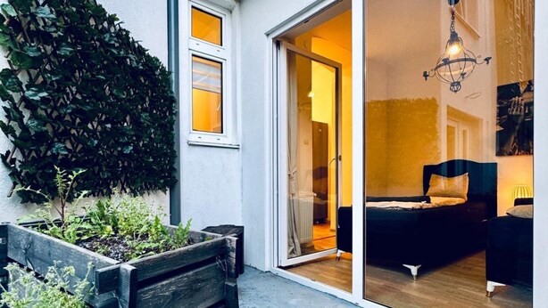 Una habitación con una cama, una silla, una jardinera de madera con plantas y una puerta corrediza de vidrio que da a otra habitación.