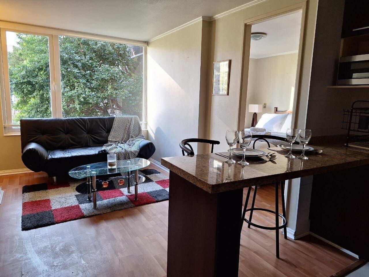 A modern kitchen with a bar area, a dining table, and a sofa in a living area with a view of trees.