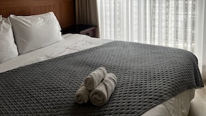 Ein Hotelzimmer mit einem großen Bett, einem Kopfteil, einem Nachttisch, einer Lampe und einem Fenster mit durchsichtigen Vorhängen.