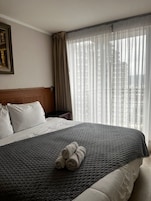 Habitación de hotel con una cama grande, cabecera, mesita de noche, lámpara y ventana con cortinas traslúcidas.