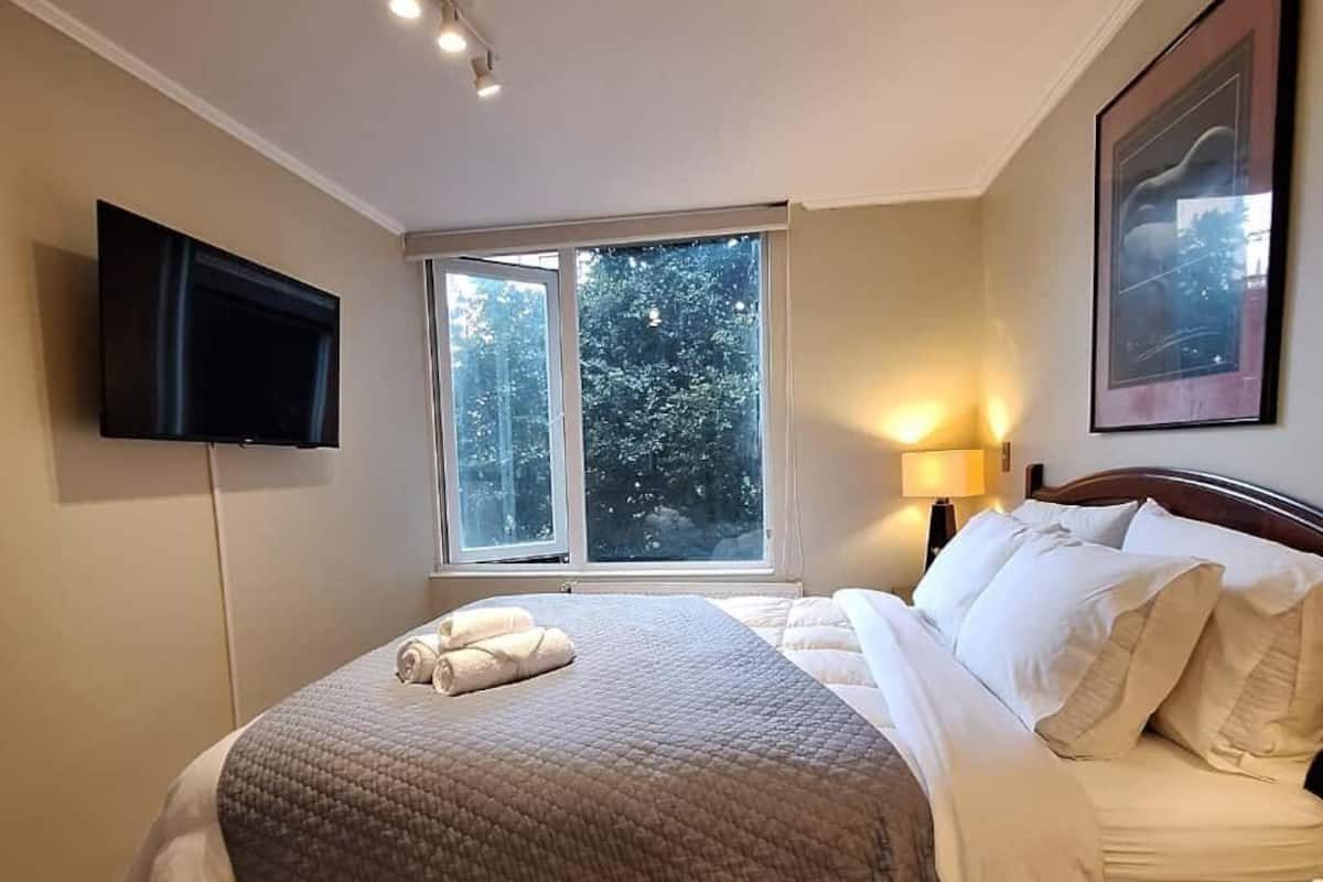 Una camera d'albergo con un letto grande, una televisione a schermo piatto fissata al muro, una finestra vista alberi e un quadro incorniciato appeso alla parete.