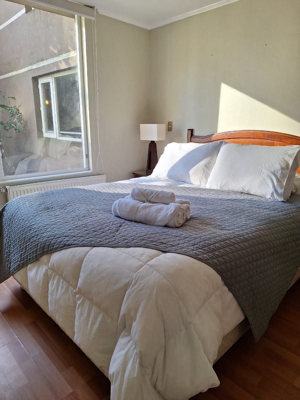 A neatly made bed with a grey quilt and white pillows, a bedside lamp, and a window with a view of the outside.
