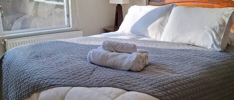 A neatly made bed with a grey quilt and white pillows, a bedside lamp, and a window with a view of the outside.