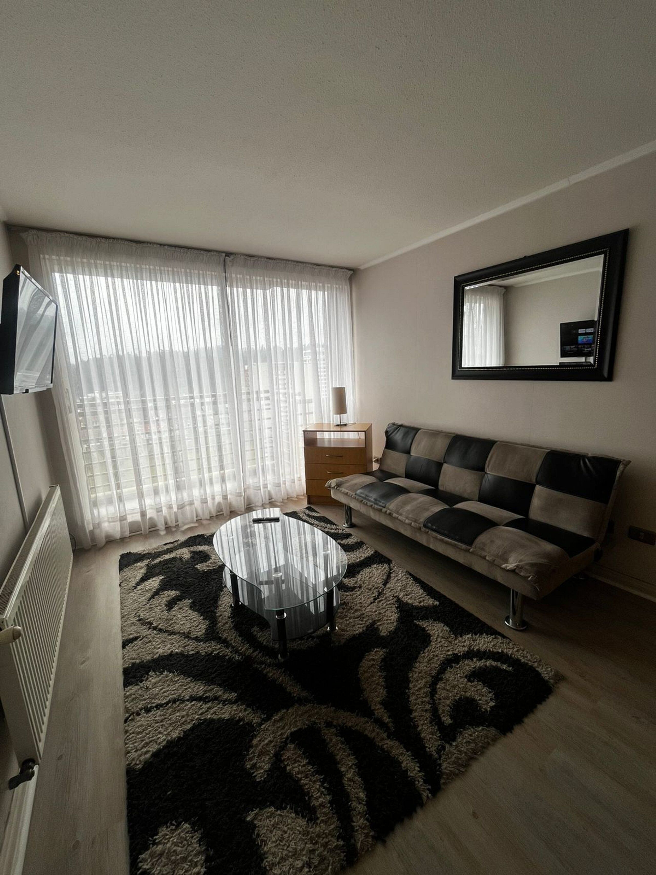 A living room with a sofa, a coffee table, a TV, and a window with sheer curtains.