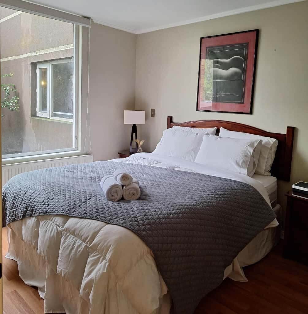Una cama bien hecha con colcha gris, almohadas blancas y toallas enrolladas sobre la cama. Hay un cuadro enmarcado en la pared encima de la cama.