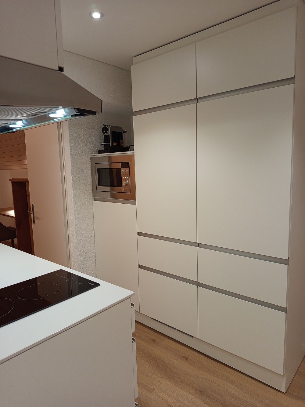 Een moderne keuken met een ingebouwde magnetron, een inductiekookplaat en een groot wit kastmeubel met verschillende vakken.