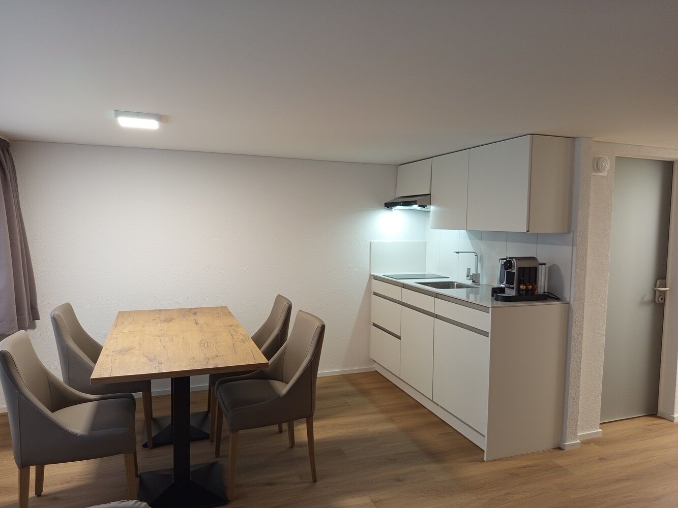 Cozinha compacta com área de refeições, apresentando mesa e cadeiras de madeira, forno embutido e máquina de café.