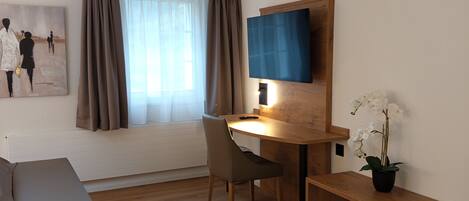 Ein modernes Wohnzimmer mit Holzboden, einem grauen Sofa, einem Couchtisch aus Holz, einem Schreibtisch mit Stuhl, einem flachbildfernseher an der Wand, einem Gemälde und einem Fenster mit Vorhängen.