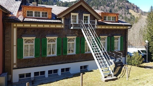 A two-story building with a wooden staircase leading to the upper floor, surrounded by mountains. - Gästehaus Rossfall 424 - Self-Check-in (Urnaesch)