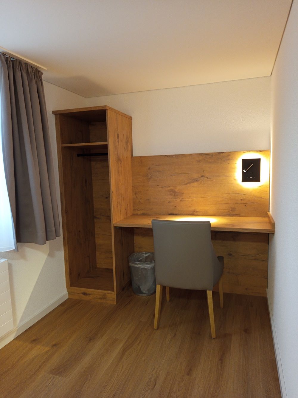 A small, compact room with a wooden desk, a chair, a built-in wardrobe, and a wall-mounted clock.