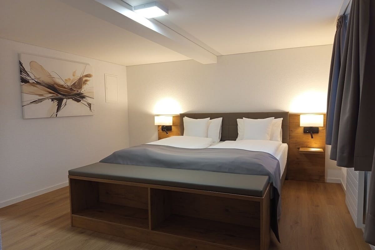 Camera d'albergo moderna con un grande letto, lampade ai comodini, una panca in legno e un'opera d'arte appesa al muro.