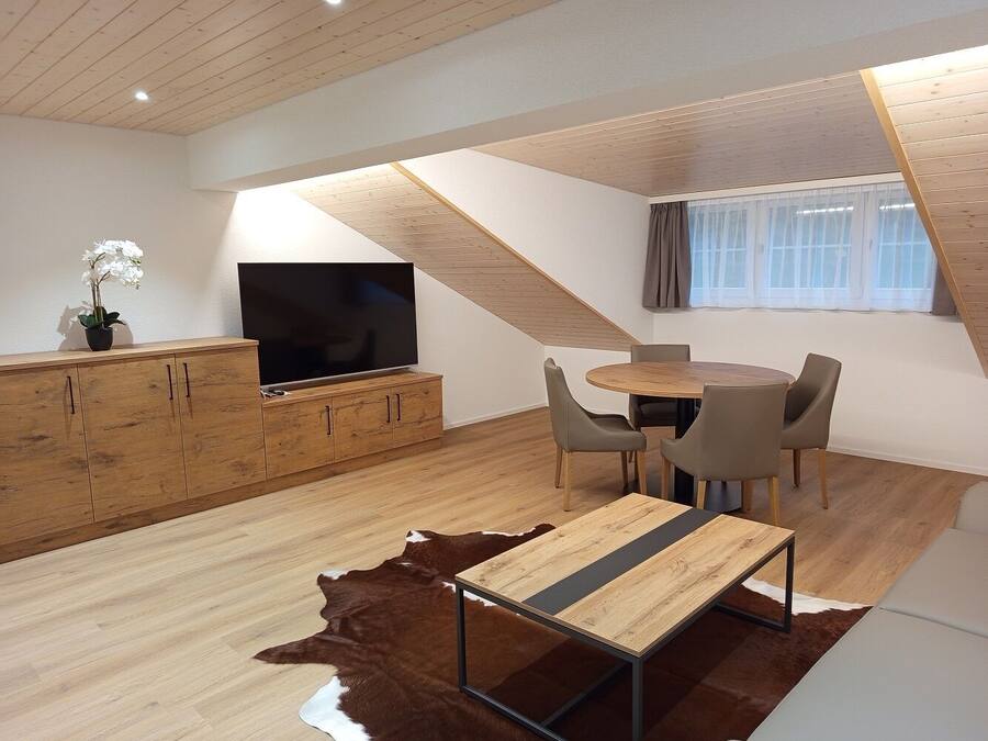 Un dormitorio tipo loft moderno con techo inclinado, piso de madera, un ventanal con cortinas translúcidas, un televisor de pantalla plana, una zona de comedor con una mesa redonda y sillas, y un armario de madera.
