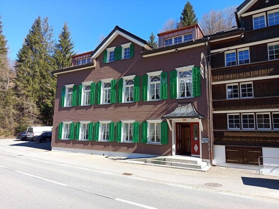 A multi-story building with a brown facade and green shutters, featuring a prominent entrance with steps leading up to it.