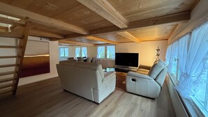 A spacious living room with a large TV, a sofa, and a recliner. There are windows with curtains and a wooden ceiling. - Gästehaus Rossfall 424 - Self-Check-in (Urnaesch)