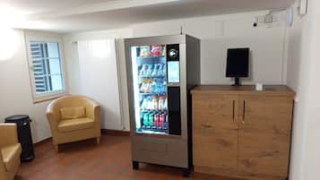 A room with a vending machine stocked with beverages, a wooden cabinet, a beige armchair, and a window with blinds.