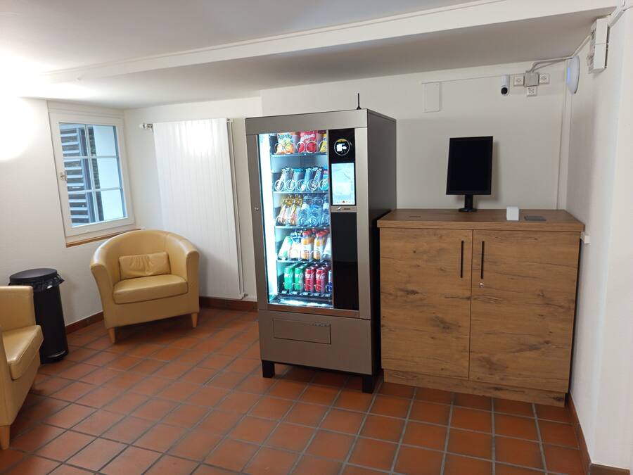 A room with a vending machine stocked with beverages, a wooden cabinet, a beige armchair, and a window with blinds.
