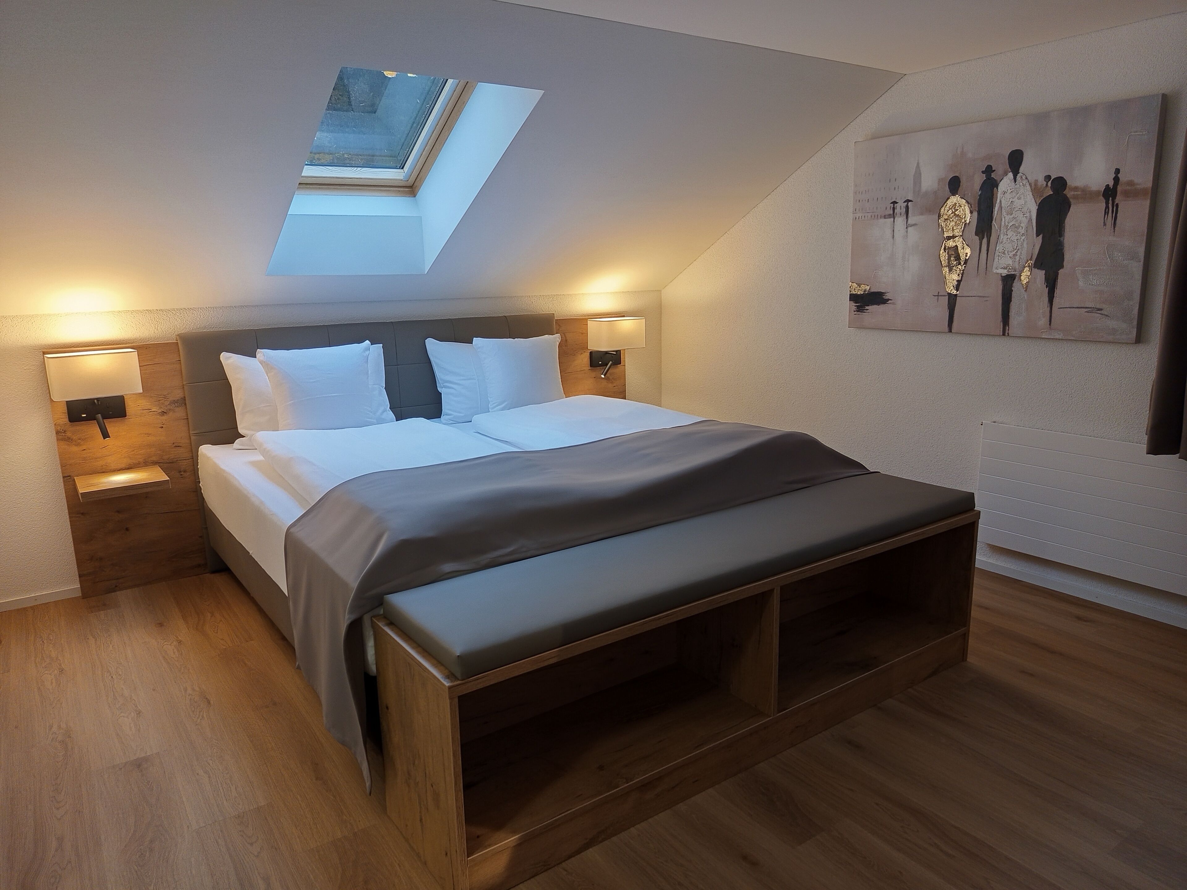 A modern bedroom with a skylight, a bed with white linens, wooden flooring, and a painting on the wall.