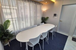 Un salón de conferencias con una larga mesa, sillas blancas y una alfombra azul. Hay plantas en macetas y una puerta con cerradura.