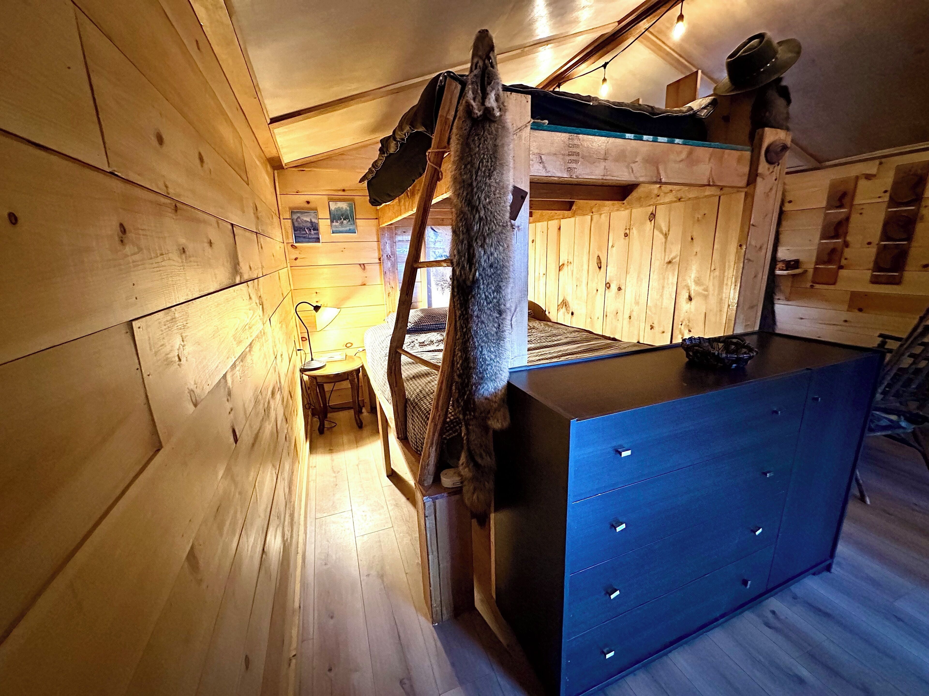 A wooden cabin with a bunk bed, a desk with a lamp, and a dresser.