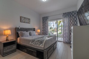 A bedroom with a large bed, two bedside tables, a nightstand, a wall-mounted lamp, and a view of the outdoors through a sliding glass door. - Apartment steps from the beach and overlooking the pool (Punta Cana)