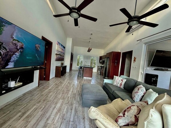 A spacious living area with a large flat-screen TV, two ceiling fans, a grey sofa, and a kitchen in the background.