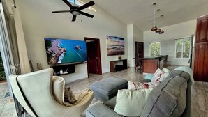 Una sala de estar moderna con un televisor de pantalla plana grande, un ventilador de techo y una zona de comedor con barra.