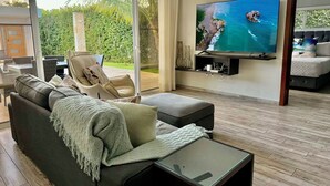 Una sala de estar moderna con un sofá, un reposapiés y una mesa de centro de vidrio. Un televisor de pantalla plana está montado en la pared, y hay un ventanal corredero que da a un espacio exterior.