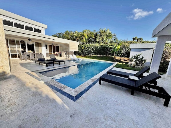 Un espace piscine extérieur moderne, doté de chaises longues, d’un coin salon abrité et d’un jardin bien entretenu.