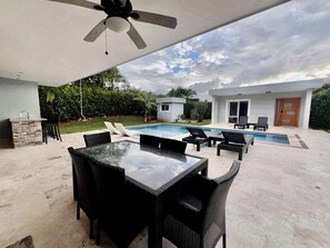 An outdoor pool area with a table and chairs, a ceiling fan, and a small structure in the background.