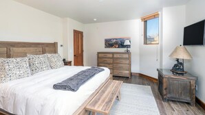 A bedroom with a large bed, wooden headboard, a bench, a dresser, a lamp, and a painting on the wall.