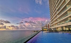 A hotel with a pool overlooking the sea, a clear sky, and a distant view of a cityscape.
