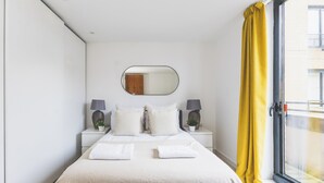 Ein modernes Schlafzimmer mit einem großen Bett, zwei Nachttischen mit Lampen, einem Spiegel über dem Bett und einem Fenster mit gelben Vorhängen.