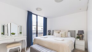 Ein Schlafzimmer mit Bett, Nachttisch, Kommode, Bank, Spiegel und einem Fenster mit Vorhängen.