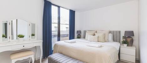 Une chambre à coucher comprenant un lit, une table de chevet, une commode, un meuble à tiroirs, un banc, un miroir et une fenêtre avec des rideaux.