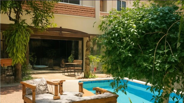 A poolside area with a wooden bench, a covered seating area, and a view of a building in the background.