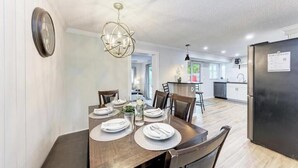 A dining area with a wooden table set for four, a modern chandelier, and a kitchen in the background. - Big backyard hangout, firepit, large primary suite (Sarasota)
