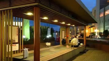 An outdoor seating area with a wooden deck and a covered section, possibly a restaurant or café, with a view of the street and buildings in the background.