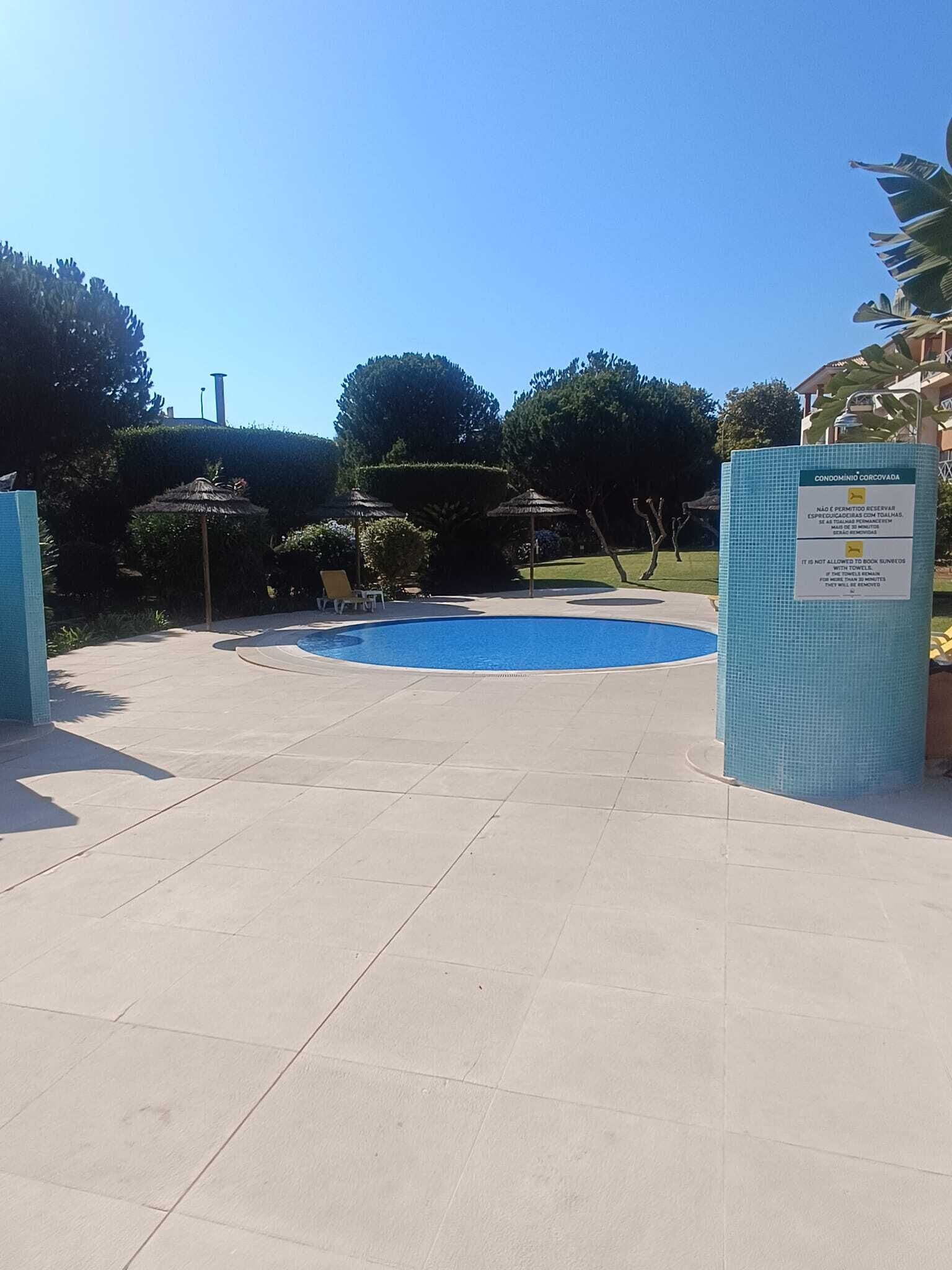 A swimming pool with a tiled deck, surrounded by greenery and a clear sky.