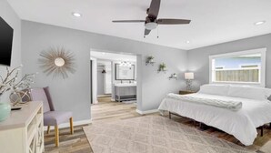 Un dormitorio con cama, ventilador de techo, mesita de noche, lámpara, repisa con plantas en la pared, y vista al baño a través de una puerta abierta.