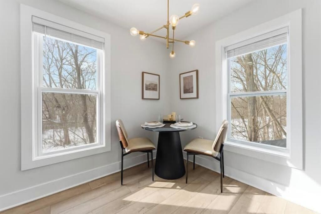 Un coin repas avec une table ronde, deux chaises, des tableaux encadrés au mur et de grandes fenêtres donnant sur des arbres sans feuilles.