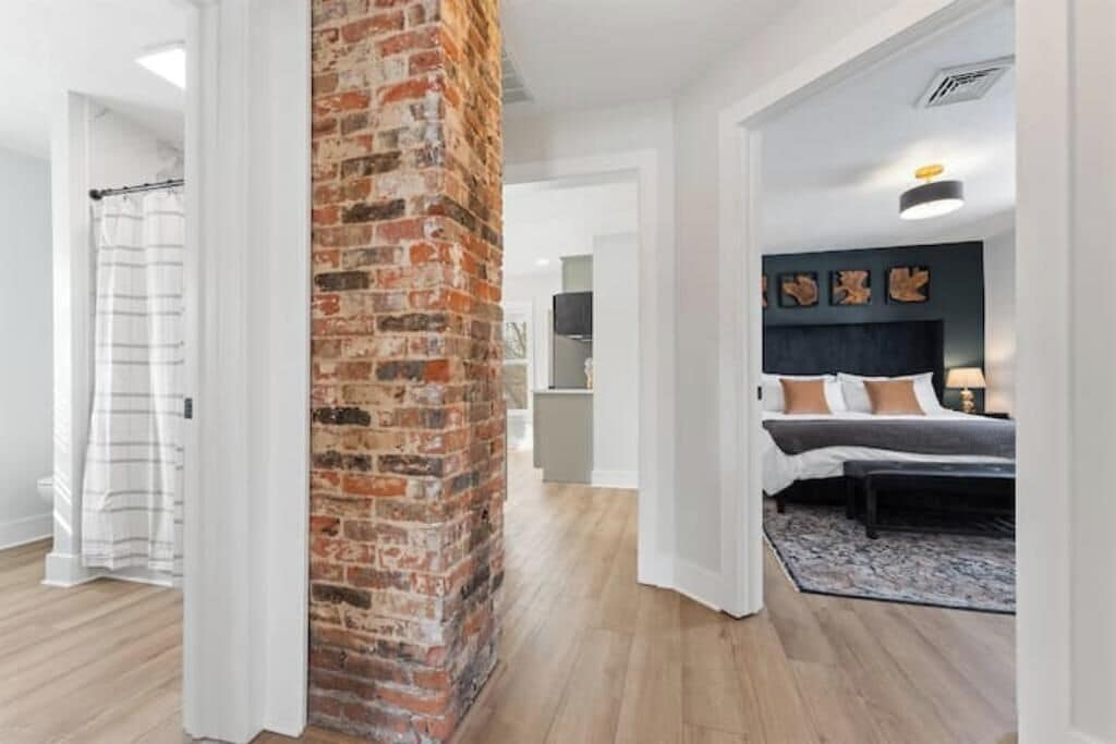 Een moderne hotelkamer met een accentmuur van baksteen, een bed met kussens, een nachtkastje en uitzicht op de gang.
