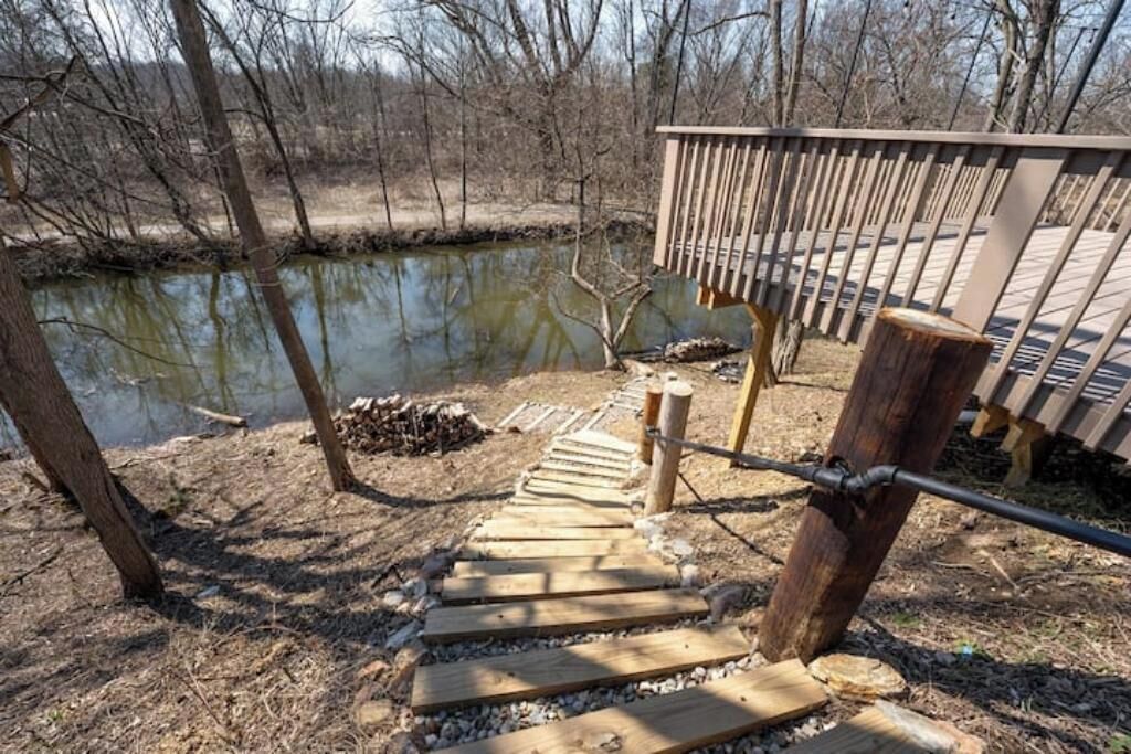 Een houten trap leidt naar een terras boven een vijver, omringd door bomen.