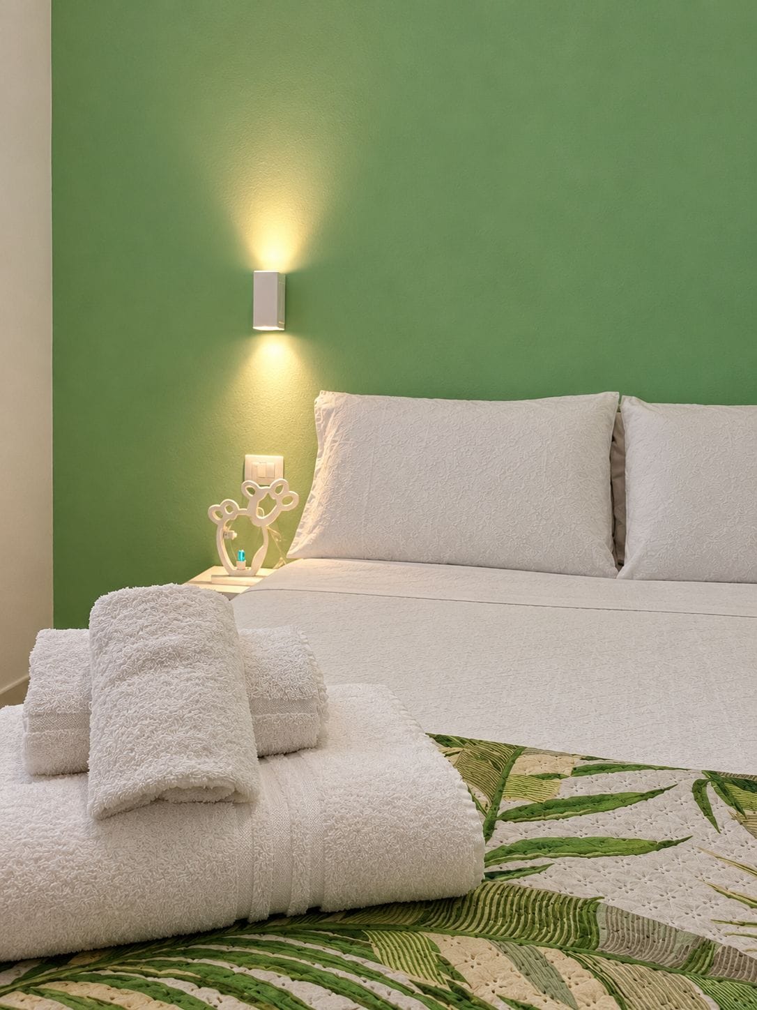Un lit bien fait, avec du linge de lit blanc, un mur vert et une table de chevet agrémentée d’une lampe et d’objets décoratifs.